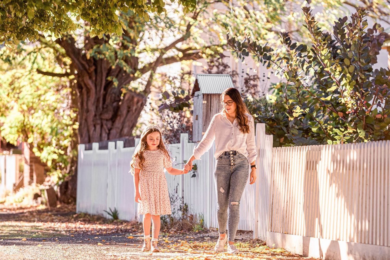 Mother and young daughter in pink dress holding hands walking along white picket fence in sunny park.
