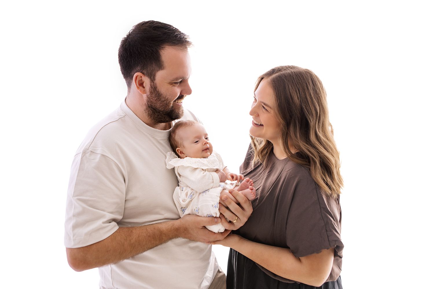 A couple smiles lovingly at their newborn baby against a white studio background.