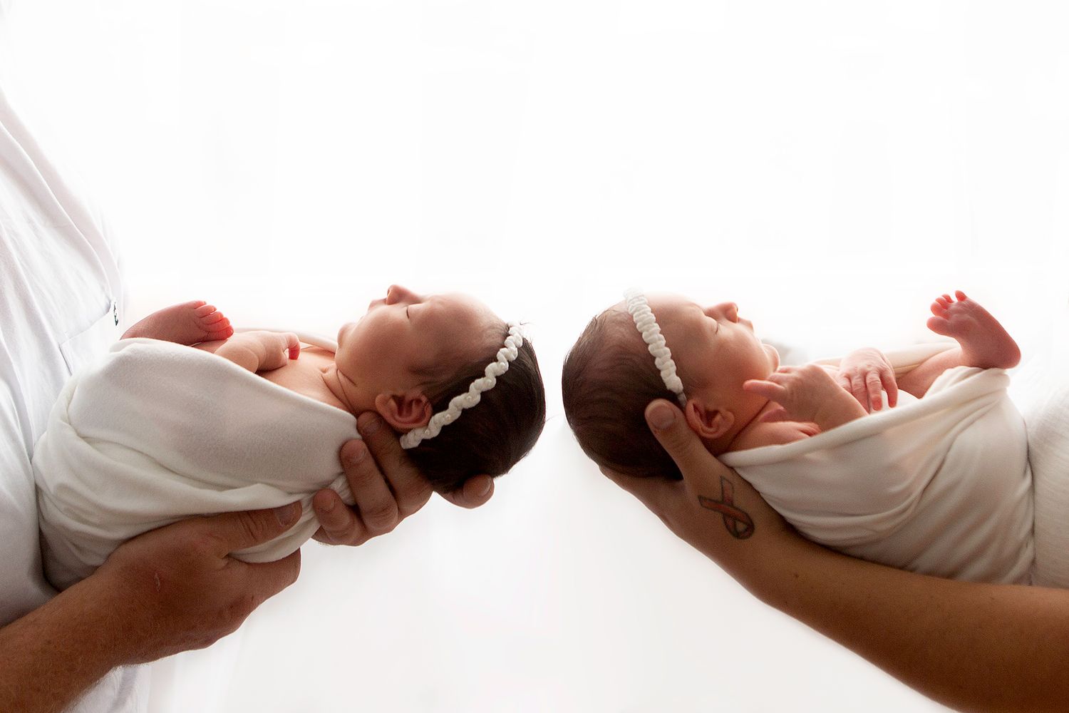 Two newborn twins swaddled in white held side by side against a bright white background.