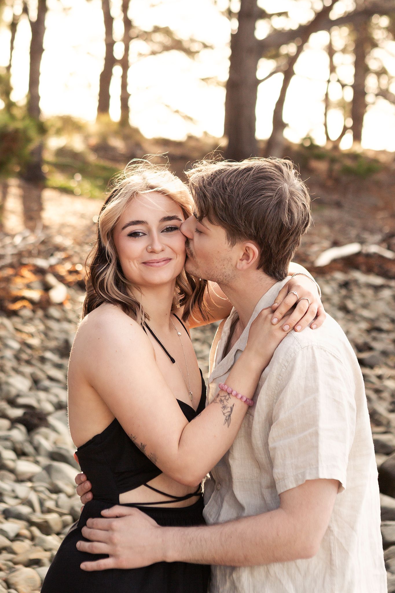 Young couple embraces on rocky beach at sunset, woman smiling as man kisses her cheek.