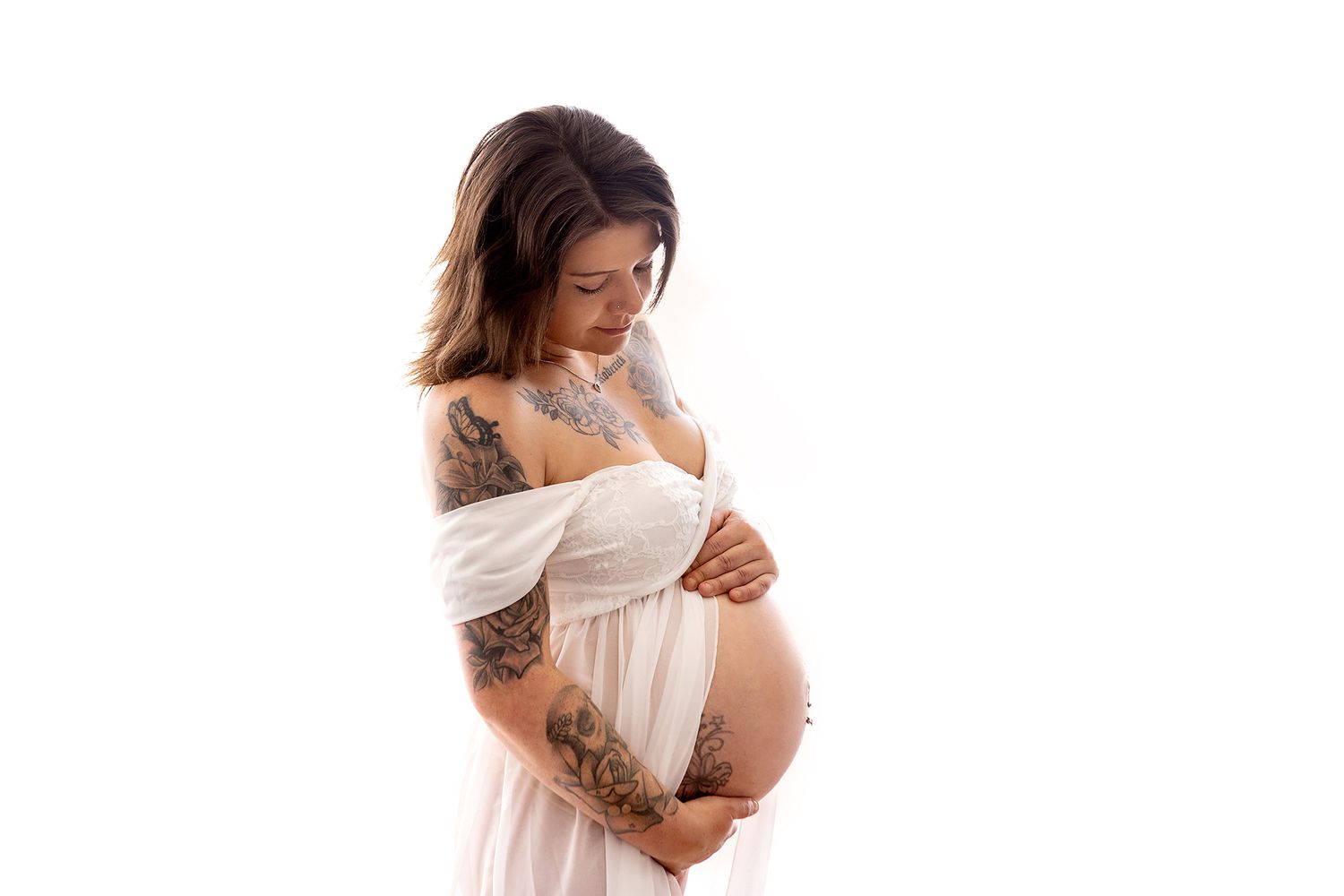 Pregnant woman with tattoos in white dress cradling baby bump on white background.