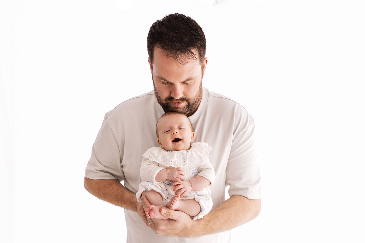 A bearded father tenderly holds his newborn baby dressed in white against a clean white background.