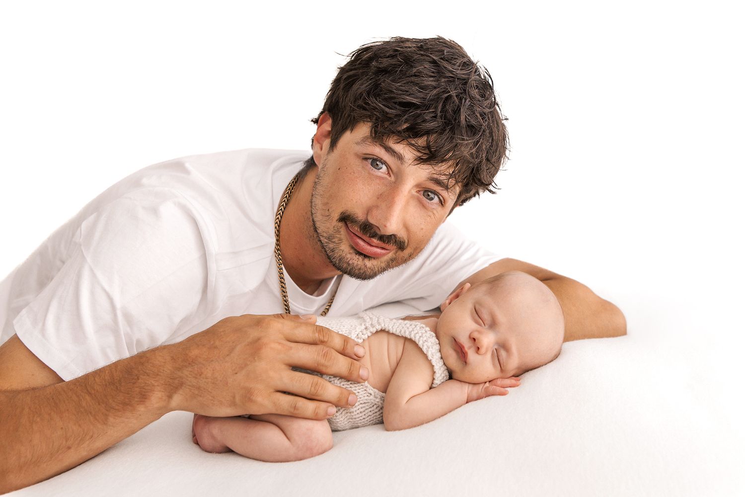 A bearded father lying beside his sleeping newborn baby on a white surface, smiling warmly.