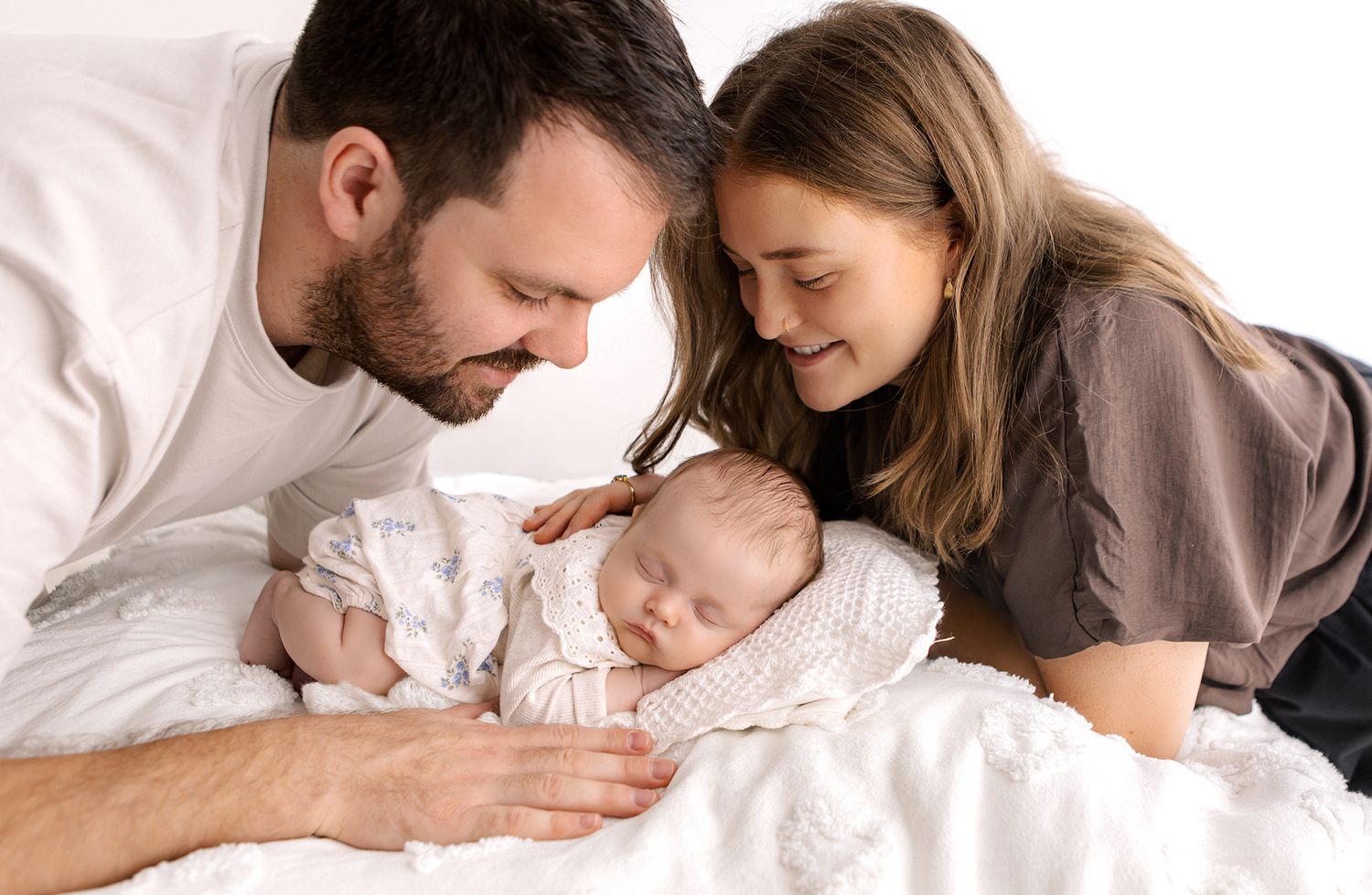 Parents lovingly gazing at their sleeping newborn baby on a white bed in a tender family portrait.