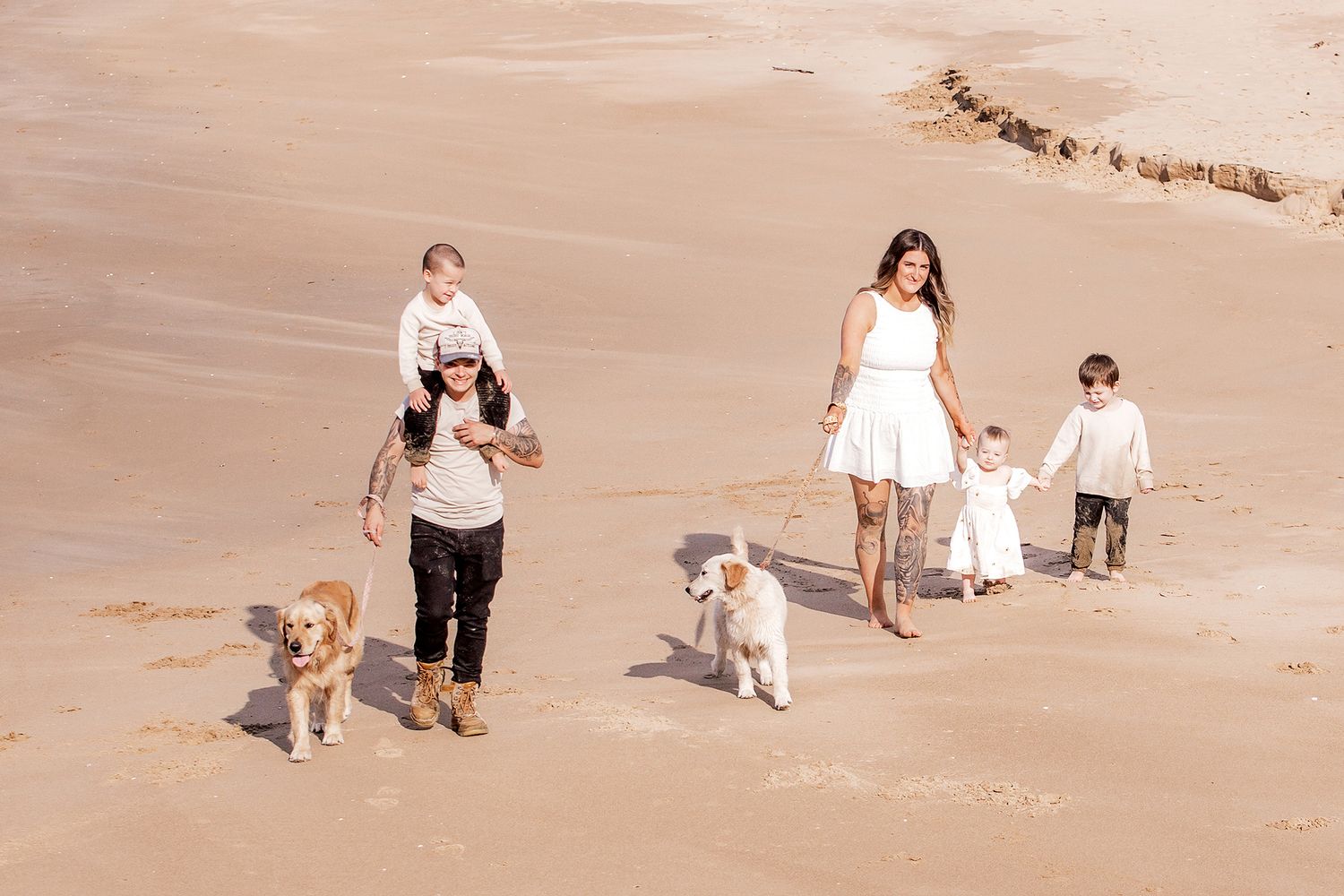 Family with two dogs walking on a sandy beach, parents with young children enjoying a sunny day outdoors.