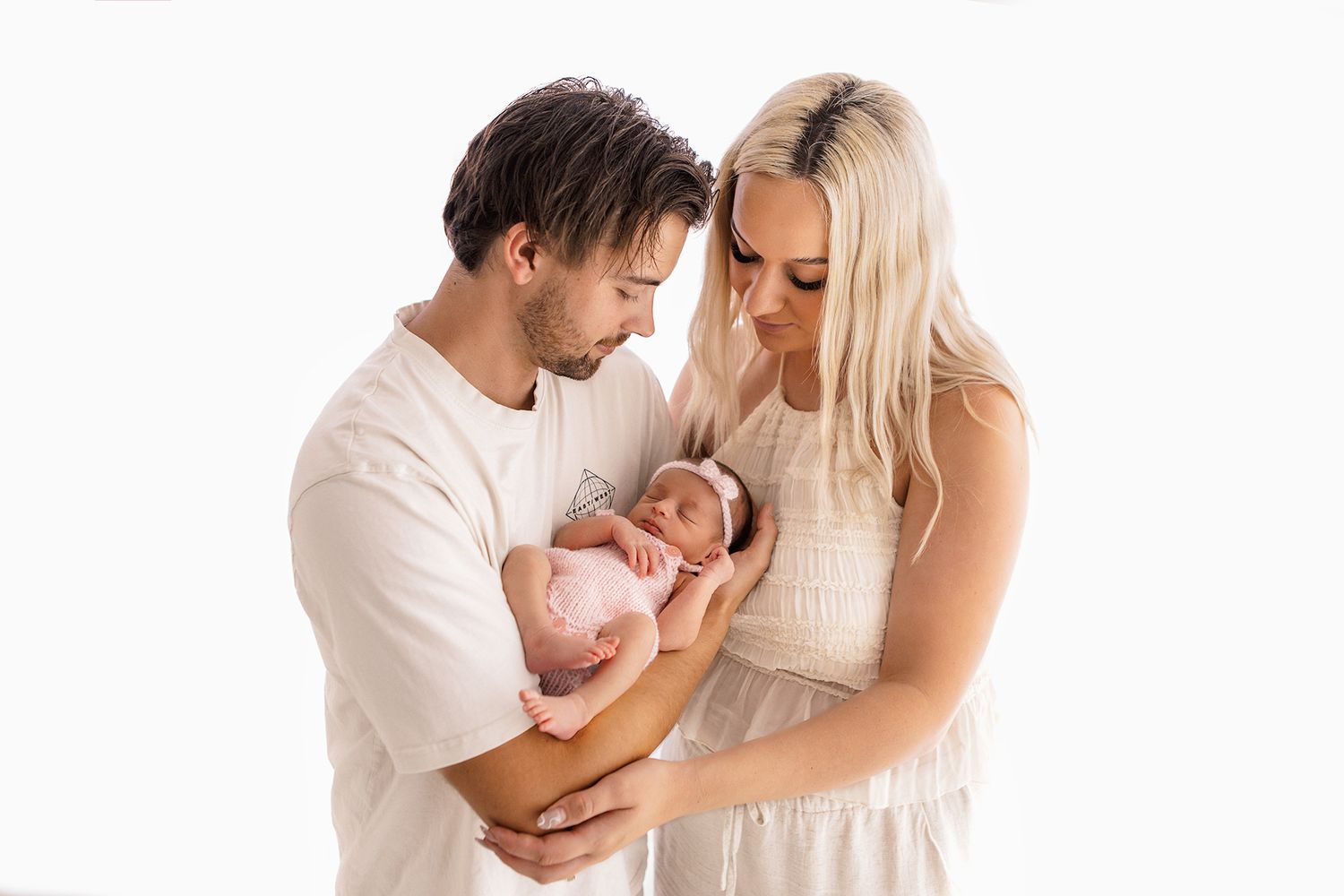 New parents tenderly hold their sleeping newborn baby in pink wrap against a white studio background.