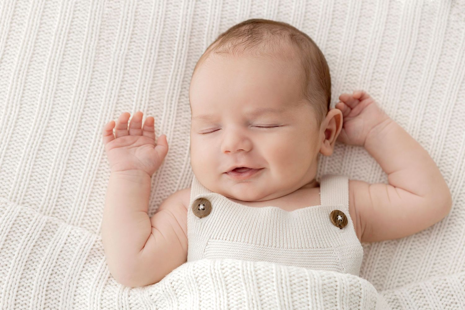 Smiling newborn baby in white knit romper lying on soft blanket with arms raised, peaceful and content.