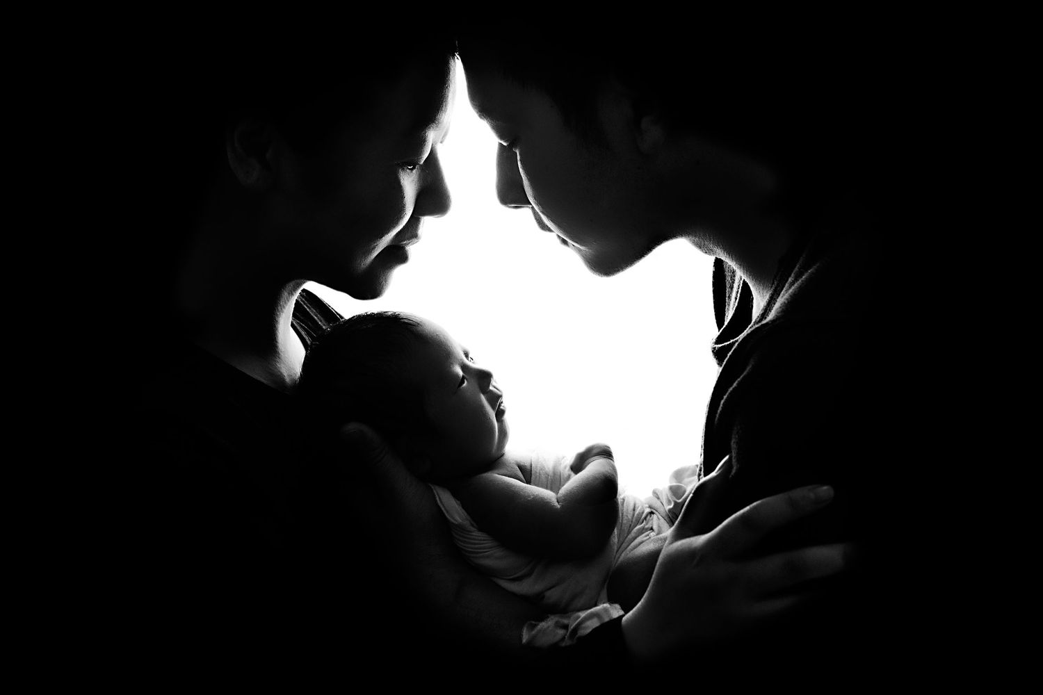 Black and white silhouette of two parents leaning in to kiss their newborn baby in a dark setting.