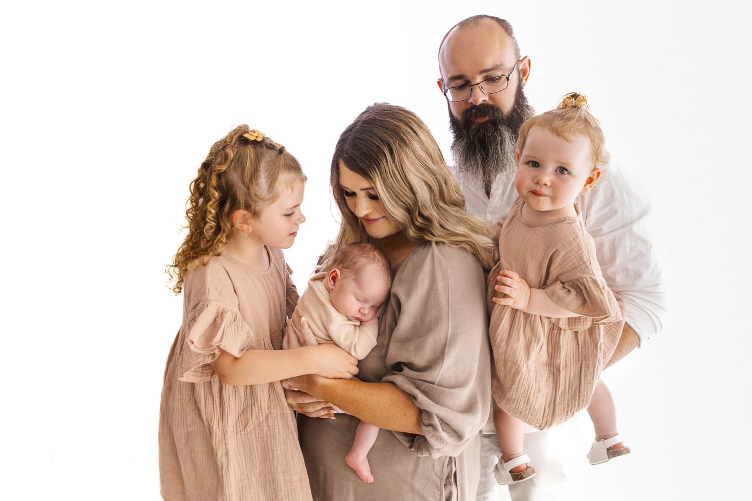 Family of five posing together, parents holding three young children, dressed in neutral tones against white background.