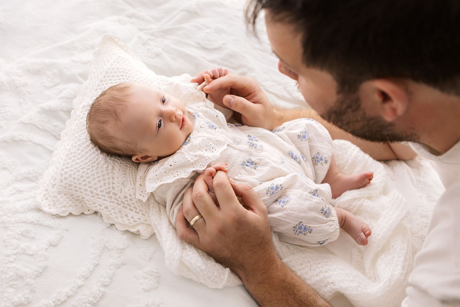 A loving father lies beside his newborn baby on a white bed, gently interacting with the infant.