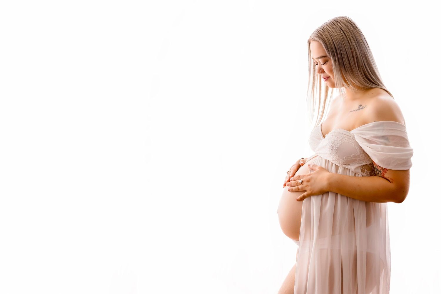 A pregnant woman in a sheer blush dress cradling her baby bump against a white background.