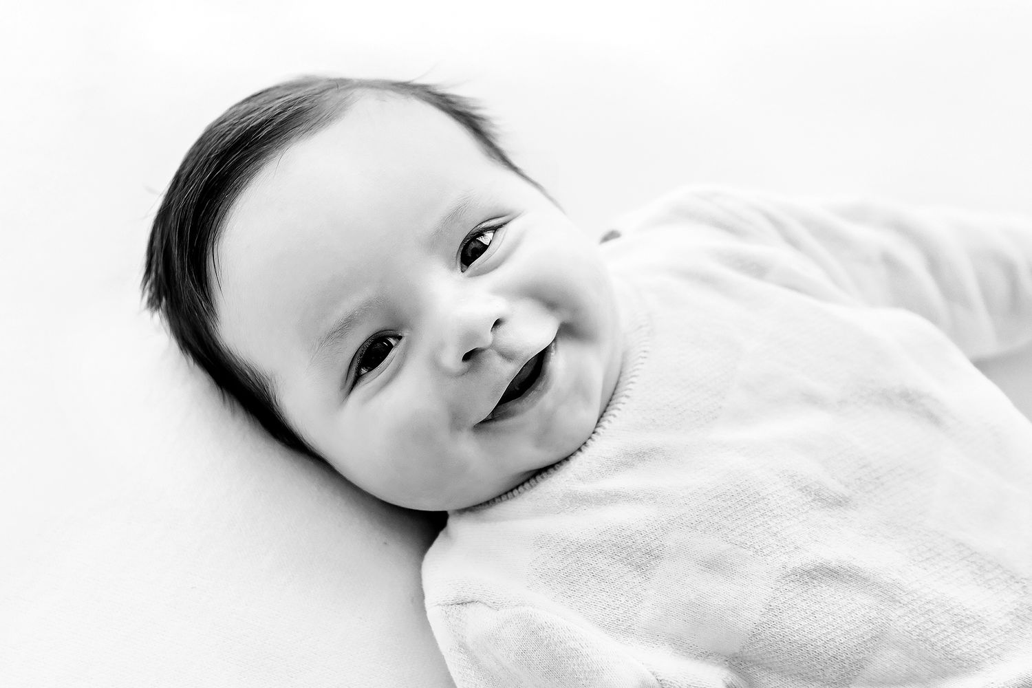 Black and white photo of a smiling newborn baby with dark hair, swaddled in a white blanket.