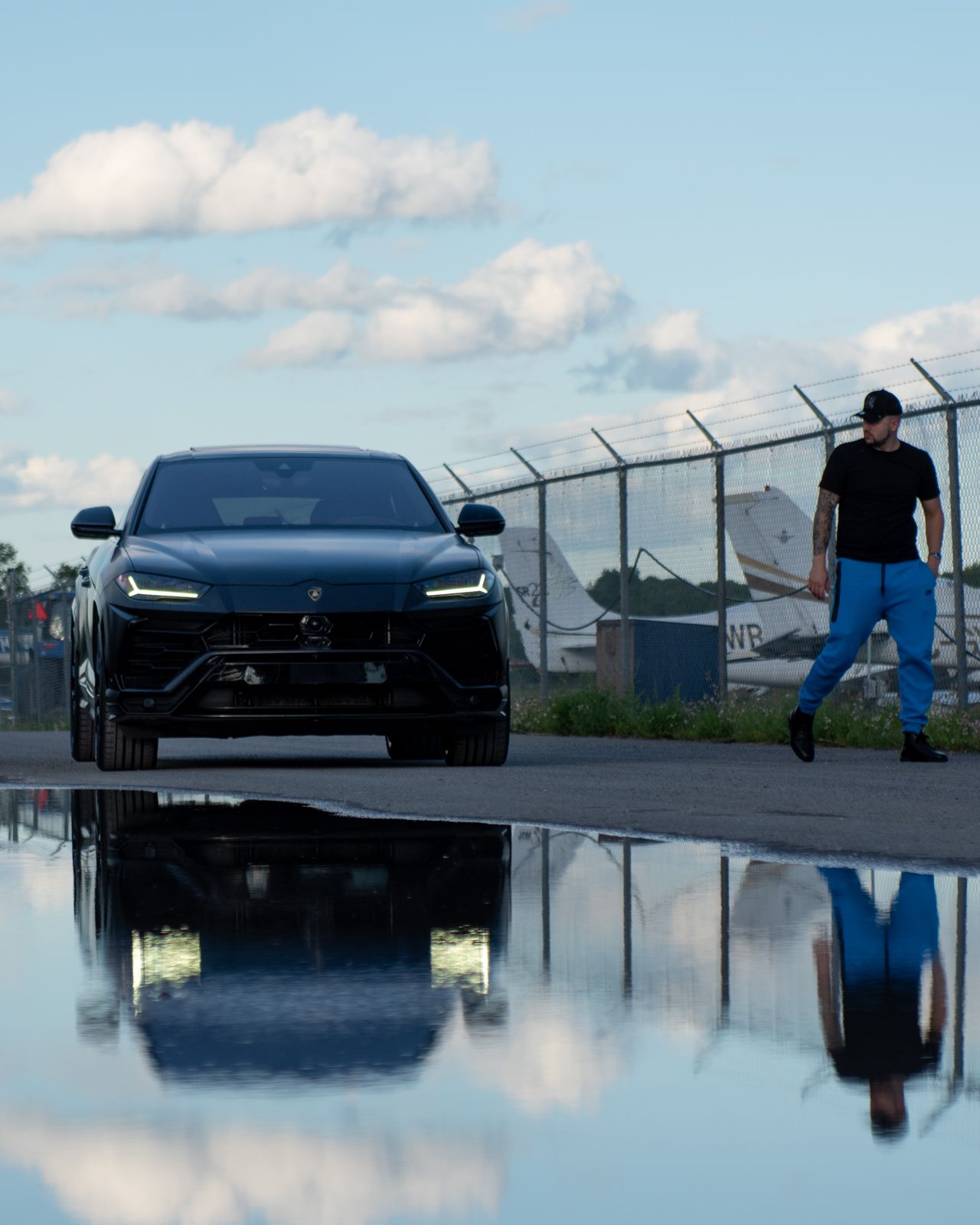 Black sports car reflected in still water on rooftop parking lot with cloudy sky in background.