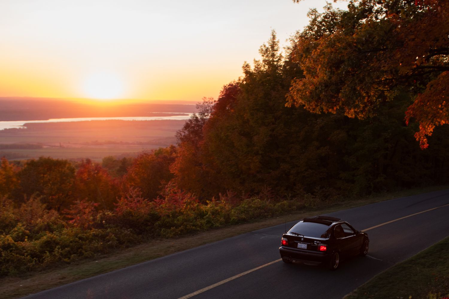 A stunning sunset illuminates a winding mountain road with silhouetted trees and distant hills in warm orange hues.