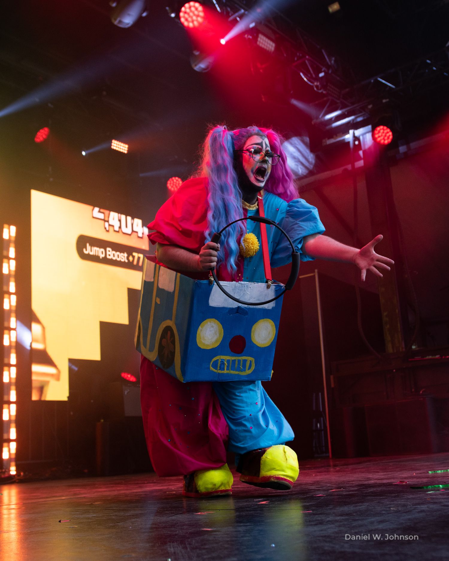 Performer in bright blue and yellow costume dances under red stage lighting.