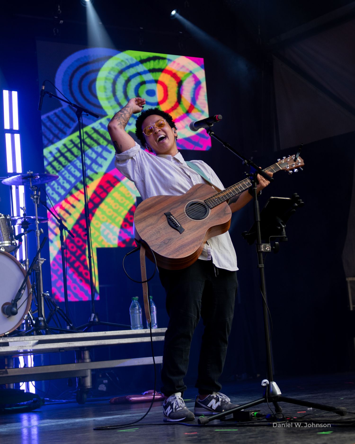 Musicians performing on stage with colorful tie-dye backdrop and acoustic guitar.