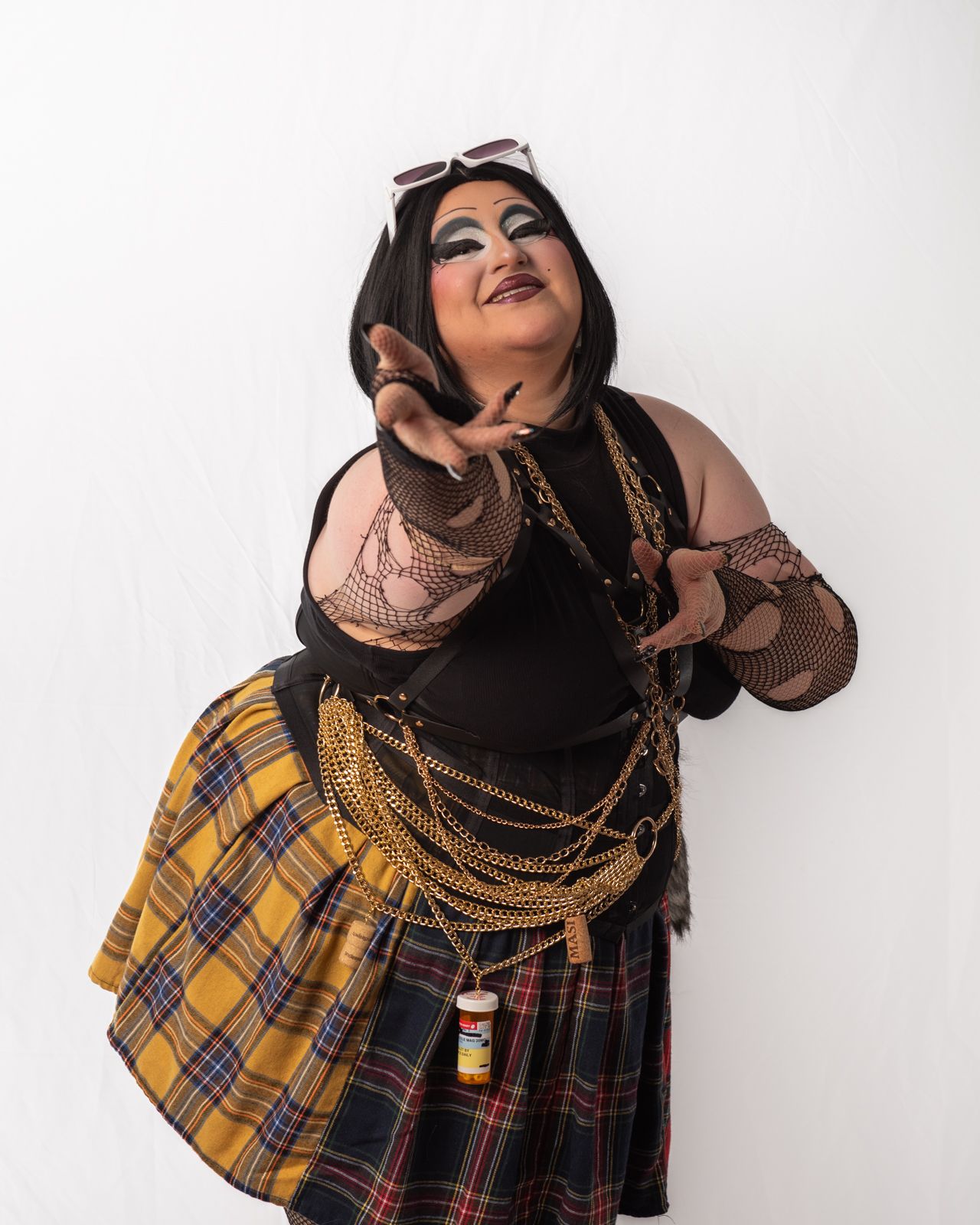 Plus size performer in plaid skirt and black corset with gold chains poses expressively against white backdrop.