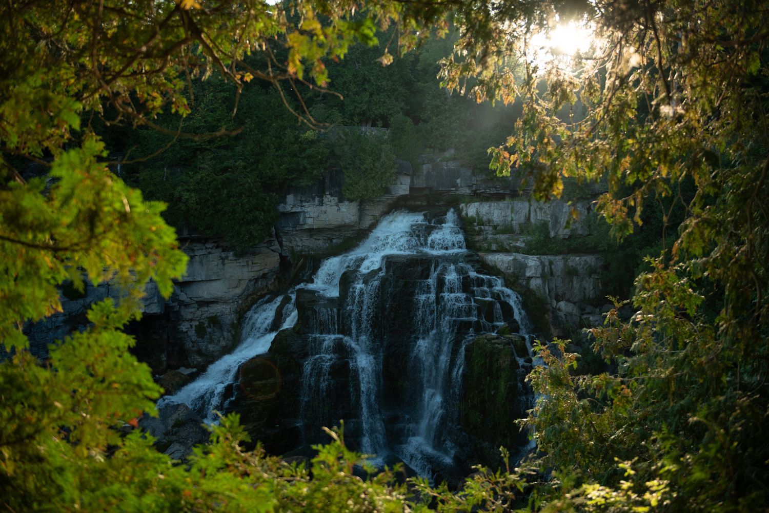 Scenic waterfall cascades down rocky cliffs surrounded by lush green foliage during golden sunset hour.