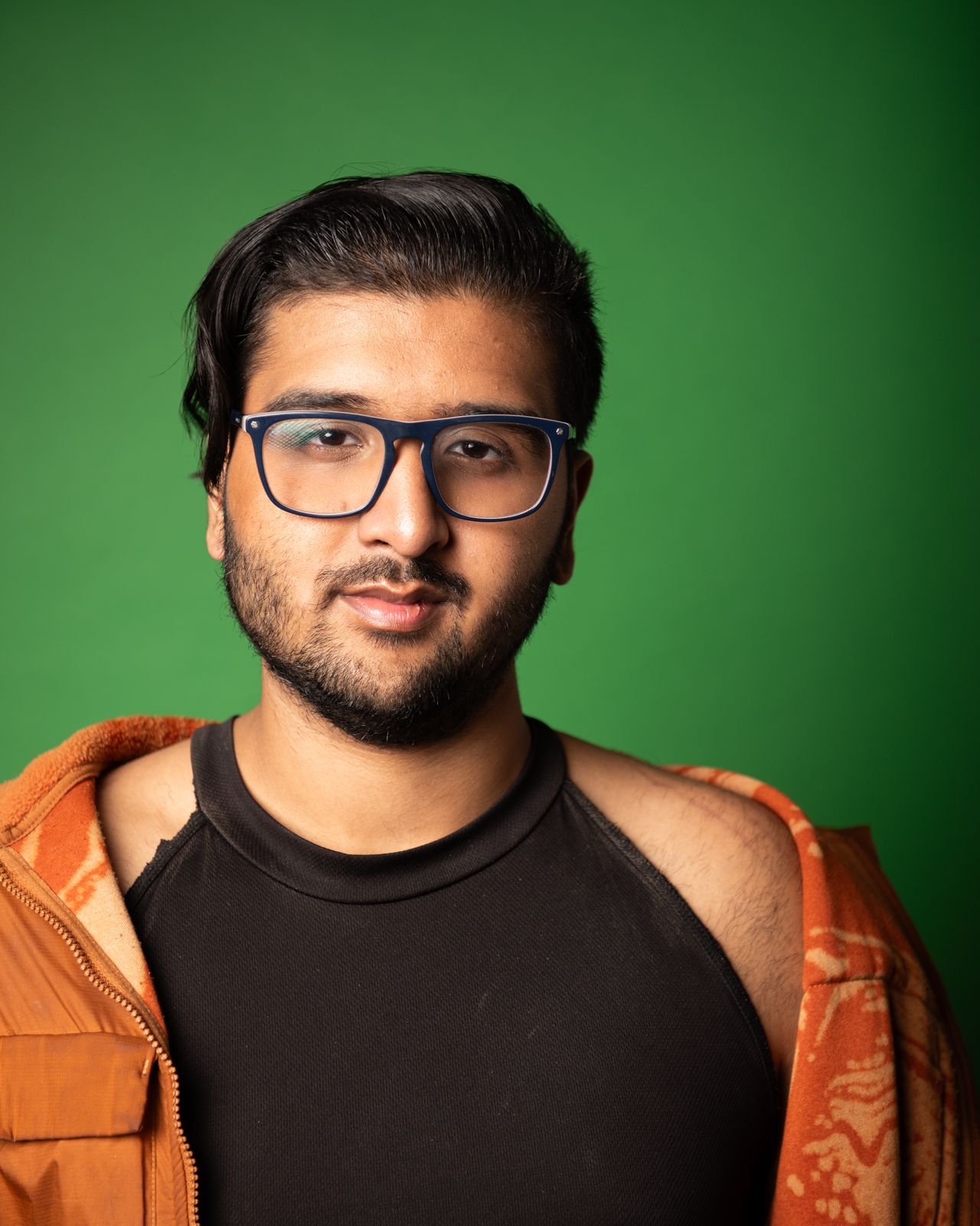 Person wearing a black shirt and orange jacket poses against a green background in a studio portrait series.