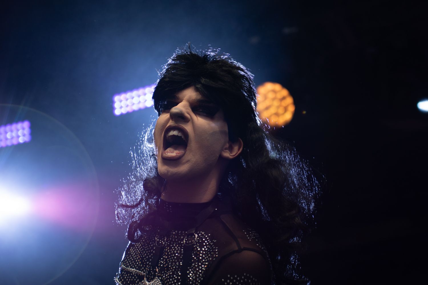 A performer in a sparkly outfit sings passionately on stage with dramatic lighting and lens flares in the background.