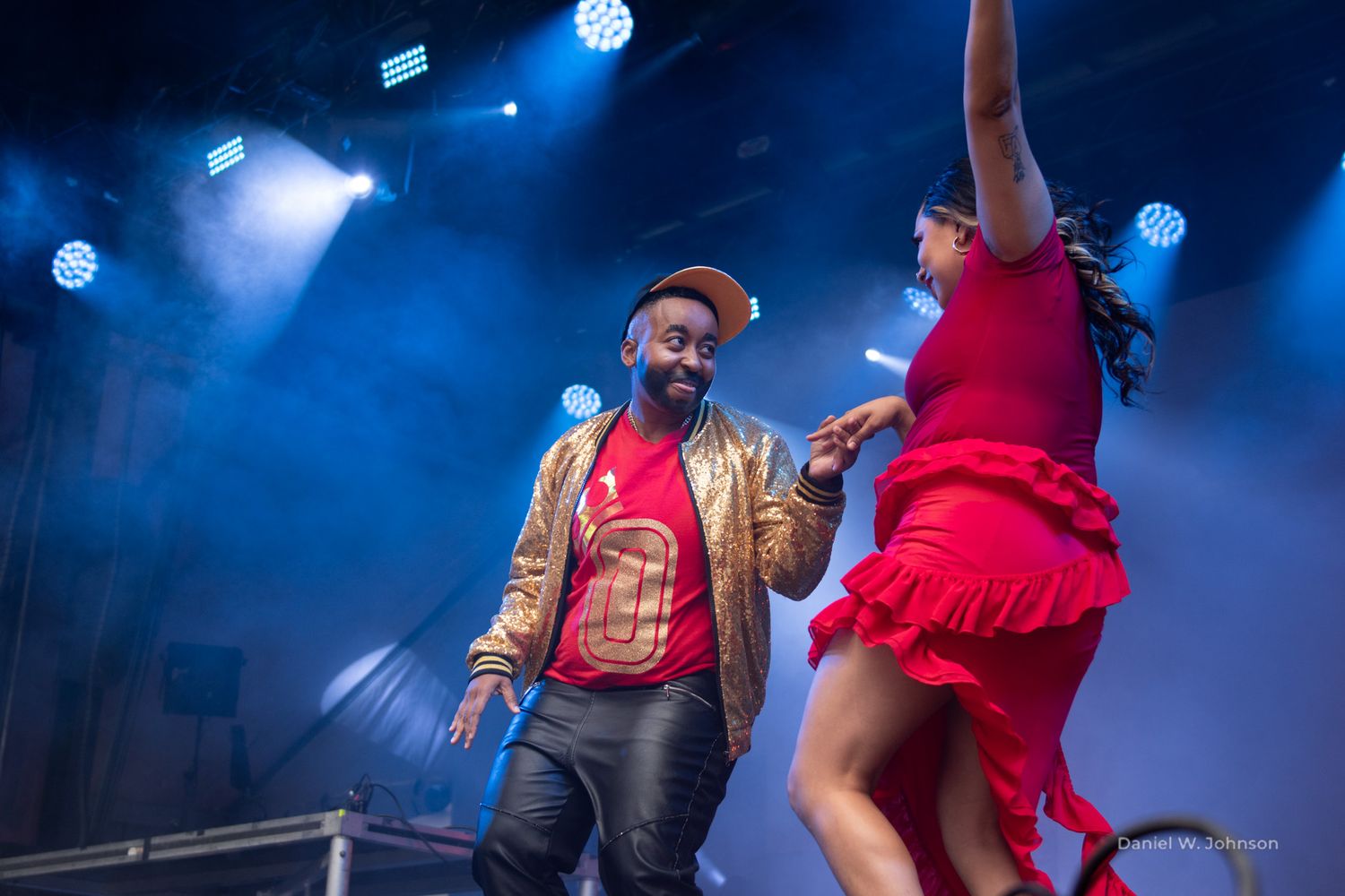 Performers in colorful costumes dance energetically on a stage with dramatic blue lighting effects.