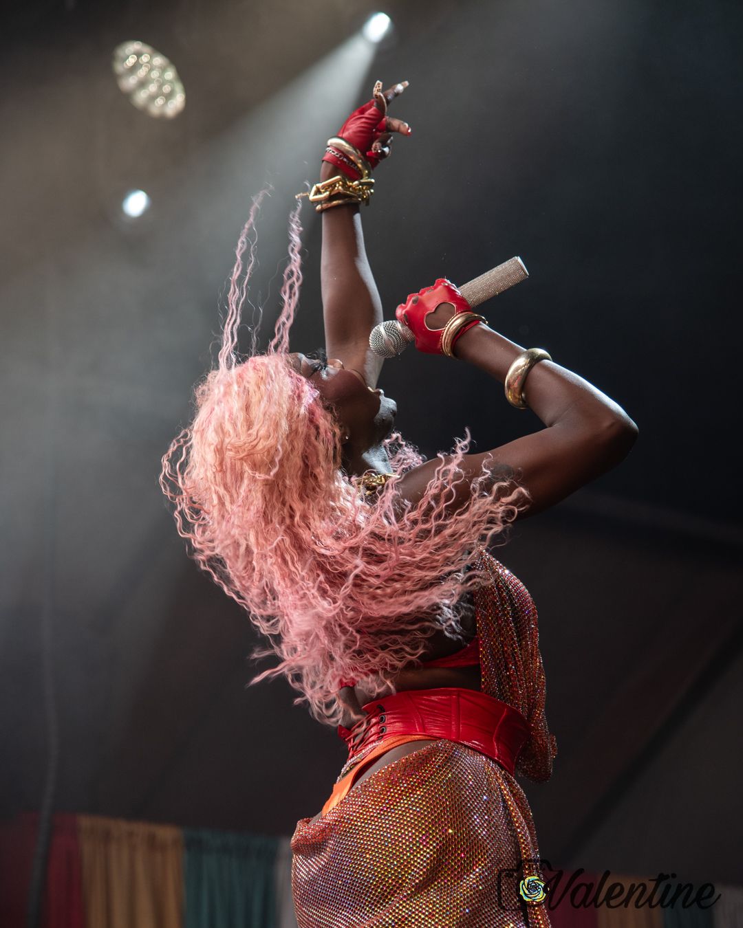 Performer in red costume with flowing pink hair dances energetically on stage during dramatic performance.