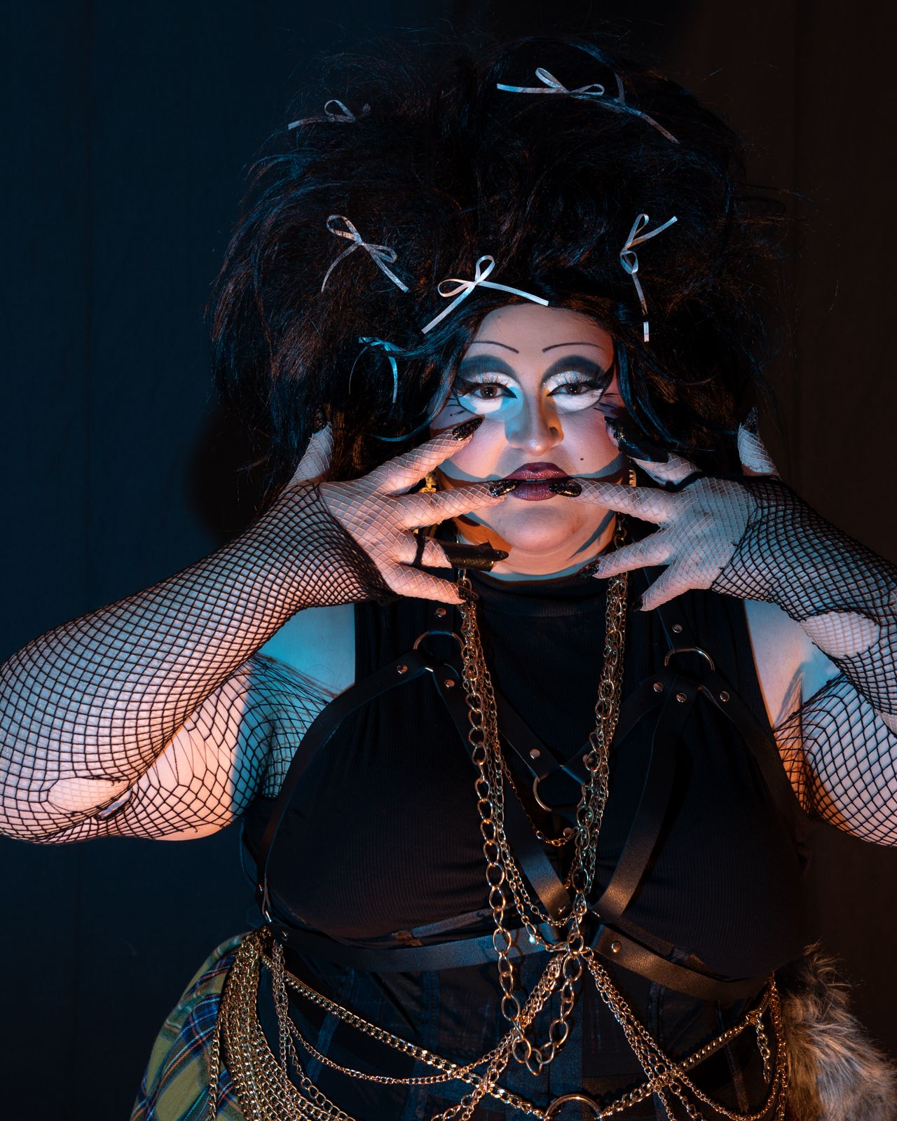 Dramatic portrait with fishnet sleeves, gold chains, and dark makeup against moody lighting in theatrical styling.