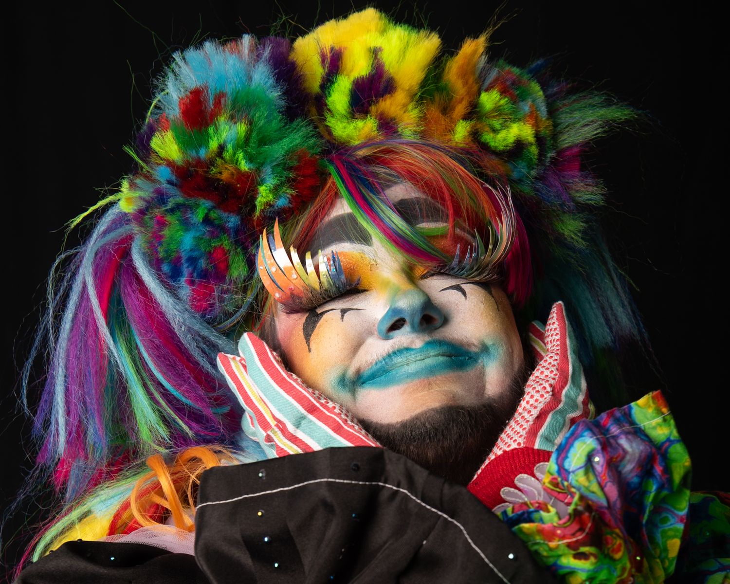 Colorful performer with rainbow hair, vivid face paint, and elaborate multicolored costume posing dramatically against dark background.