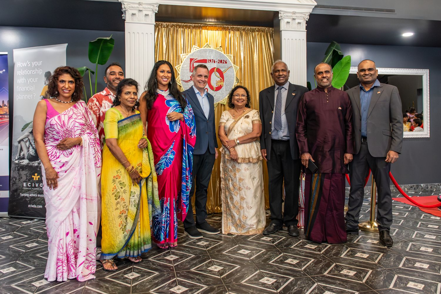 Group of elegantly dressed people posing together at a formal cultural event, wearing traditional South Asian attire and western suits.