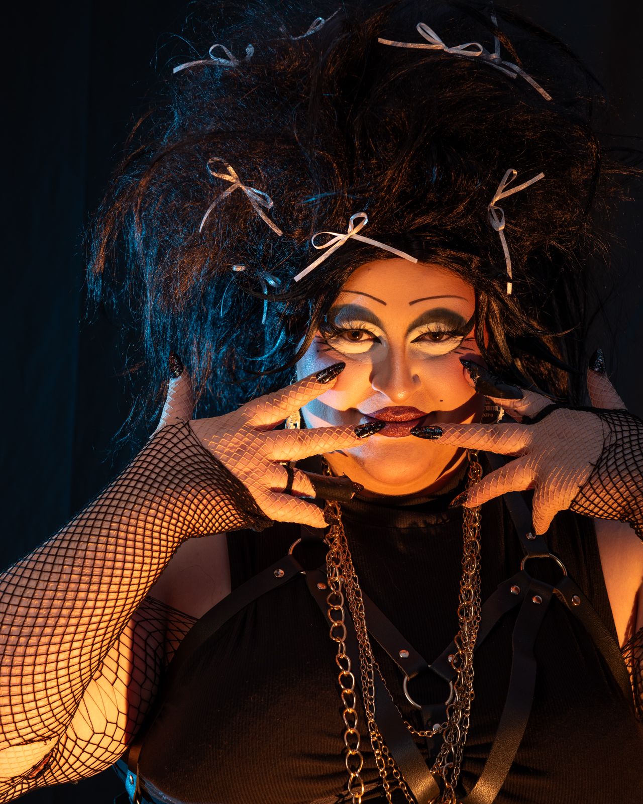 Dramatic portrait with bold eye makeup, wild dark hair with bows, fishnet sleeves and chains against dark background.