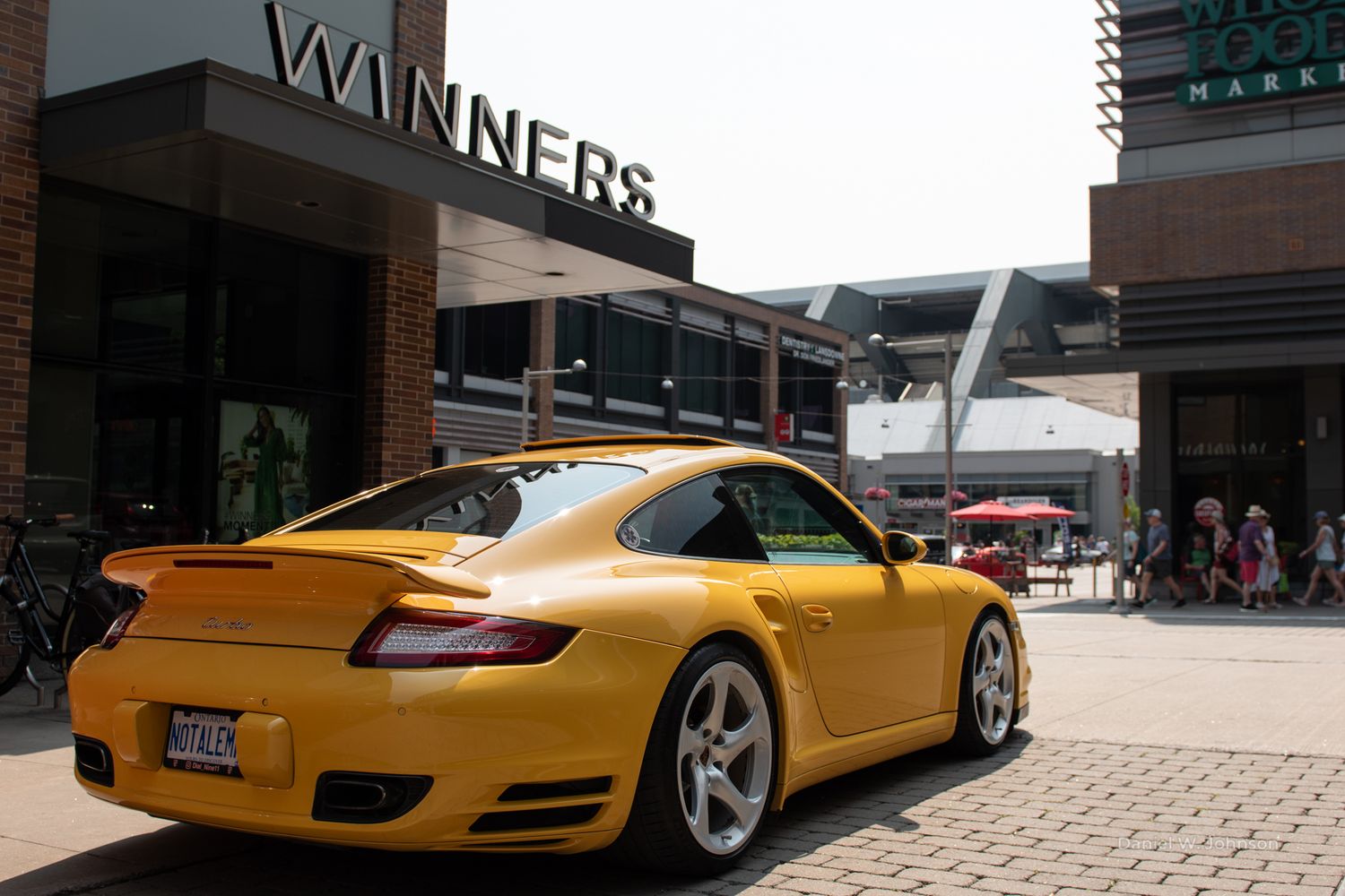 A yellow Porsche 911 GT3 sports car parked on a brick paved surface near retail storefronts.