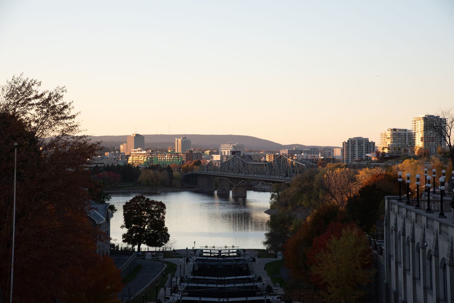 A scenic cityscape view at sunset showing buildings along a waterfront with mountains in the background.