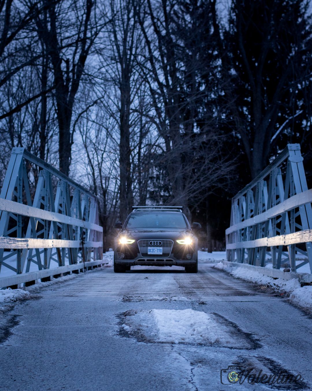 An Audi car crossing a snowy white bridge at dusk with headlights illuminating the path ahead.