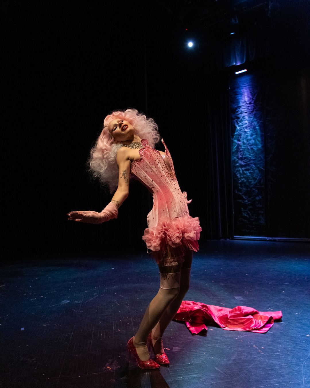 Performer in pink dress and blonde wig strikes dramatic pose under blue stage lighting.