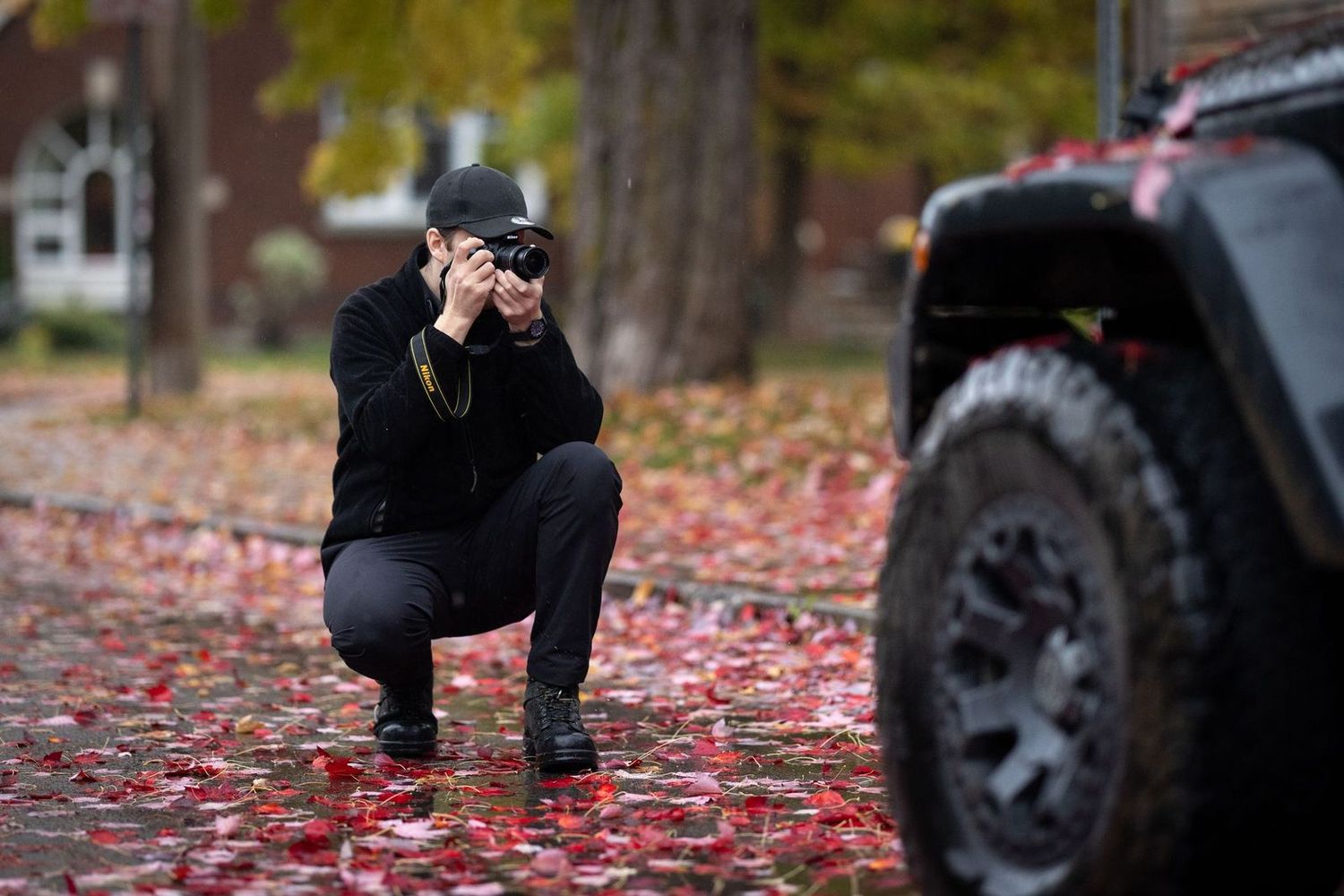 Photographer crouching to take photos among fallen autumn leaves next to a black off-road vehicle.
