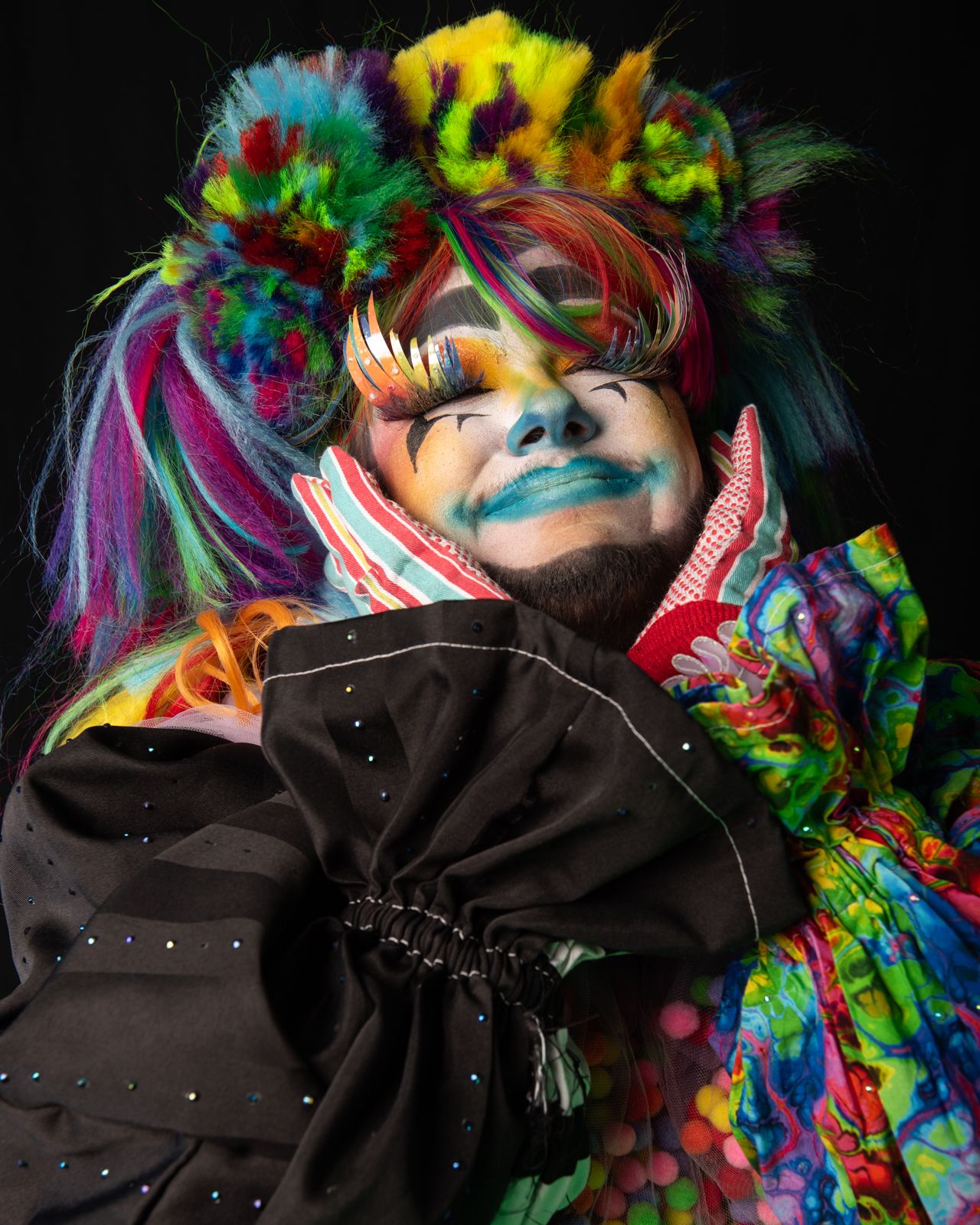 Artistic portrait featuring vibrant rainbow colored hair and feathers against a black background in a creative fashion style.