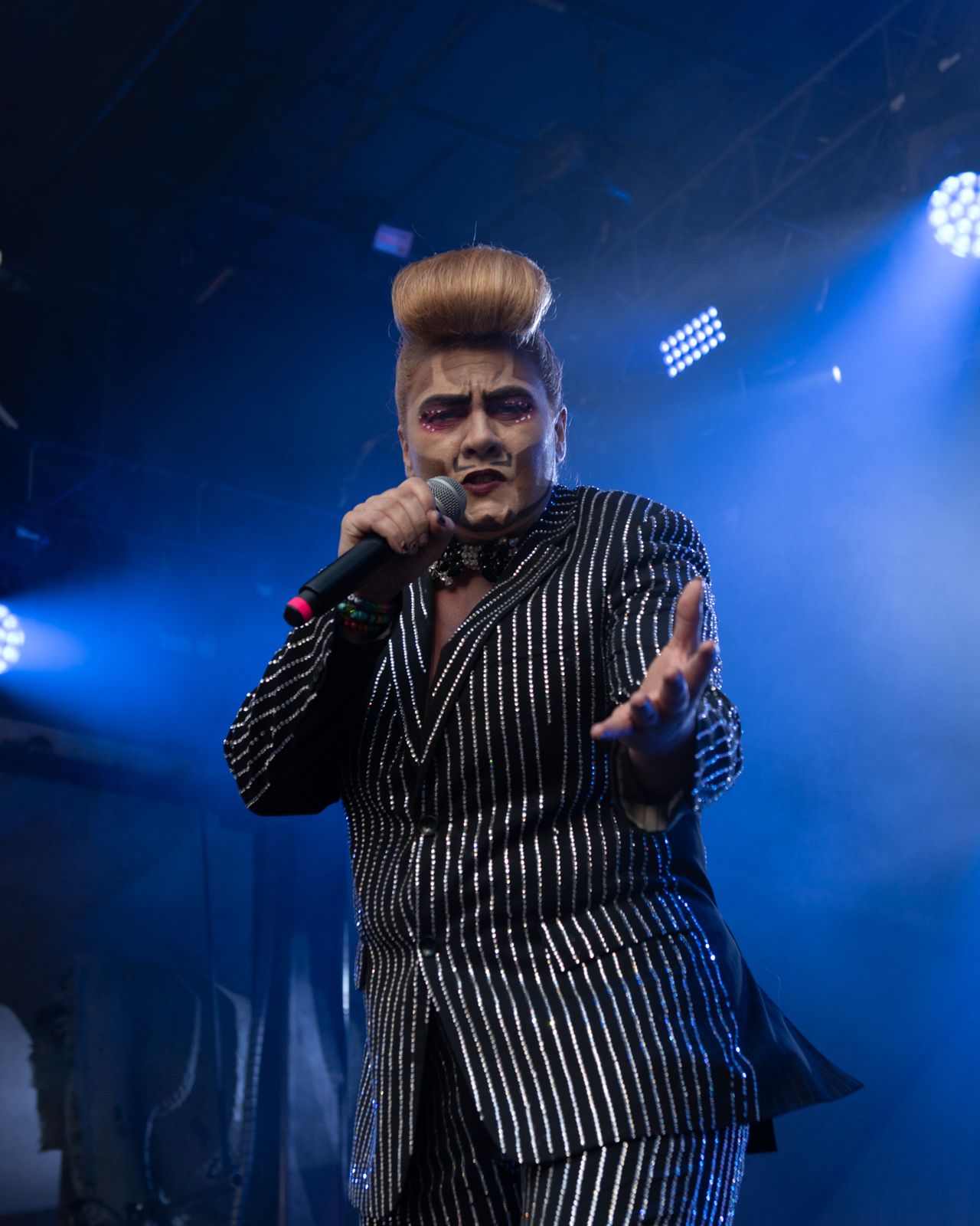 A performer in a striped suit jacket gestures energetically on a dark stage illuminated by dramatic blue lighting.