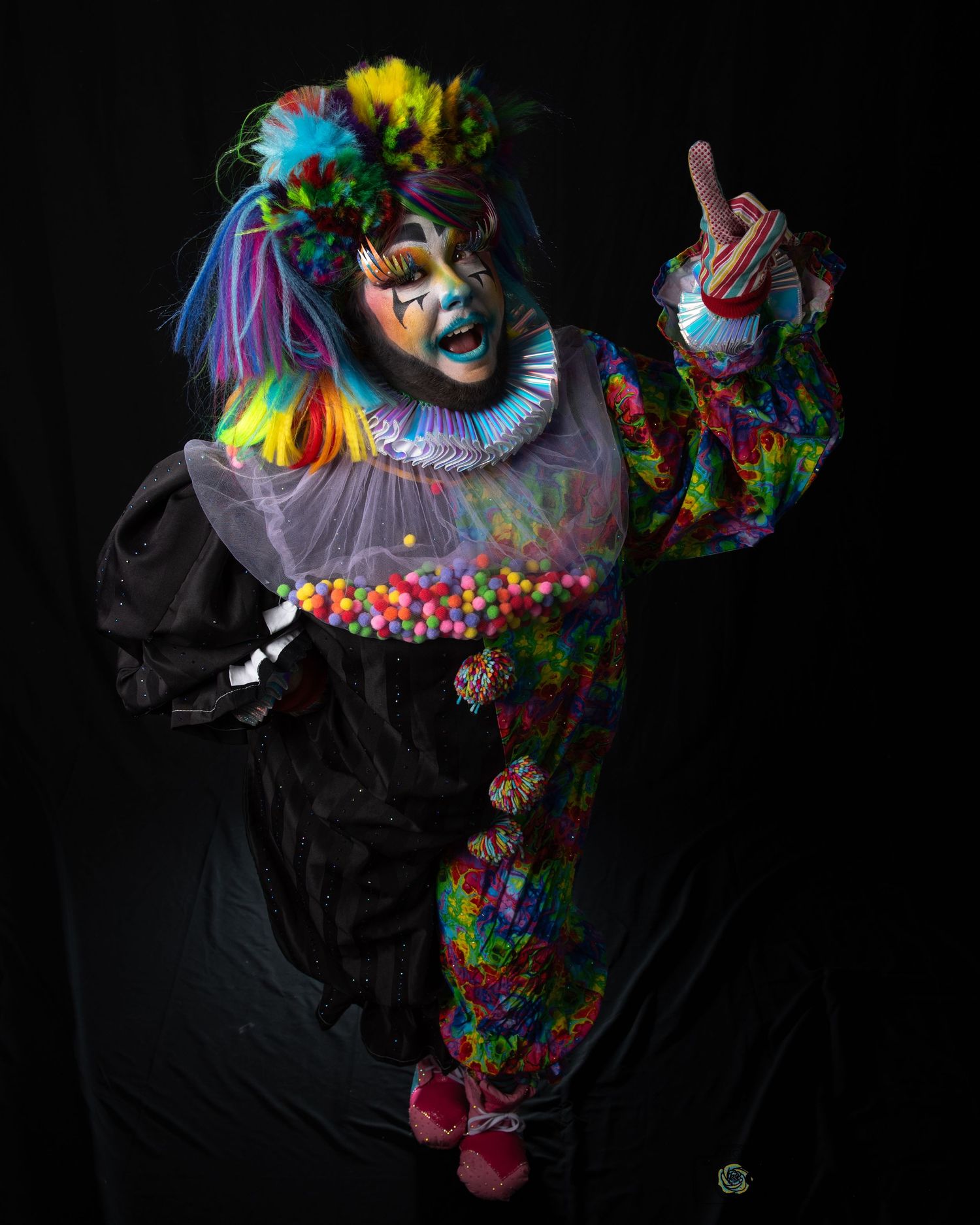 Colorful clown costume with rainbow hair and ruffled collar against dark background in dramatic studio lighting.