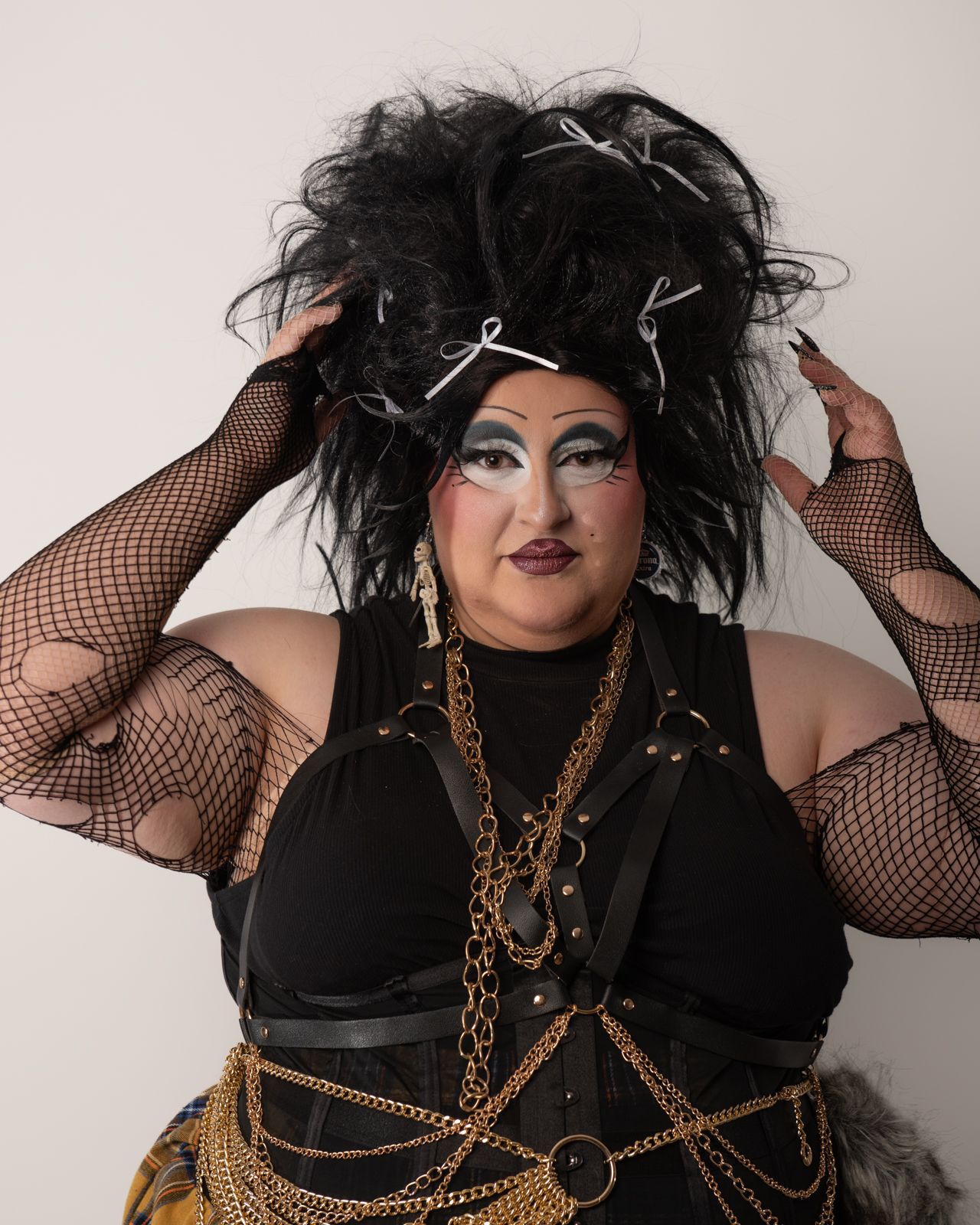 Drag performer in dramatic black hairstyle wearing fishnet sleeves and gold chain harness poses confidently.