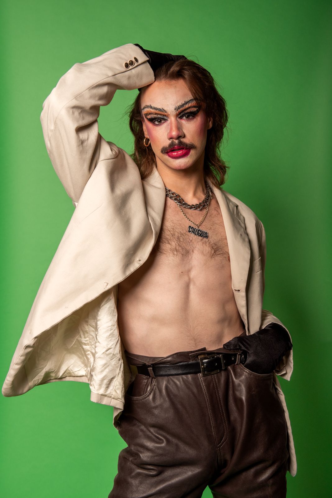 A fashion model poses in a cream blazer and brown leather dress against a bright green background.