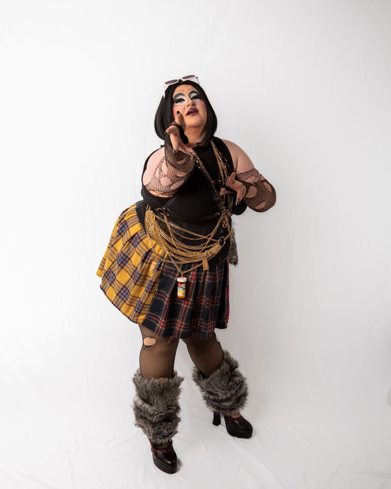 A person in a steampunk-inspired costume with plaid skirt, fur boots, and leather accessories poses dramatically against white.