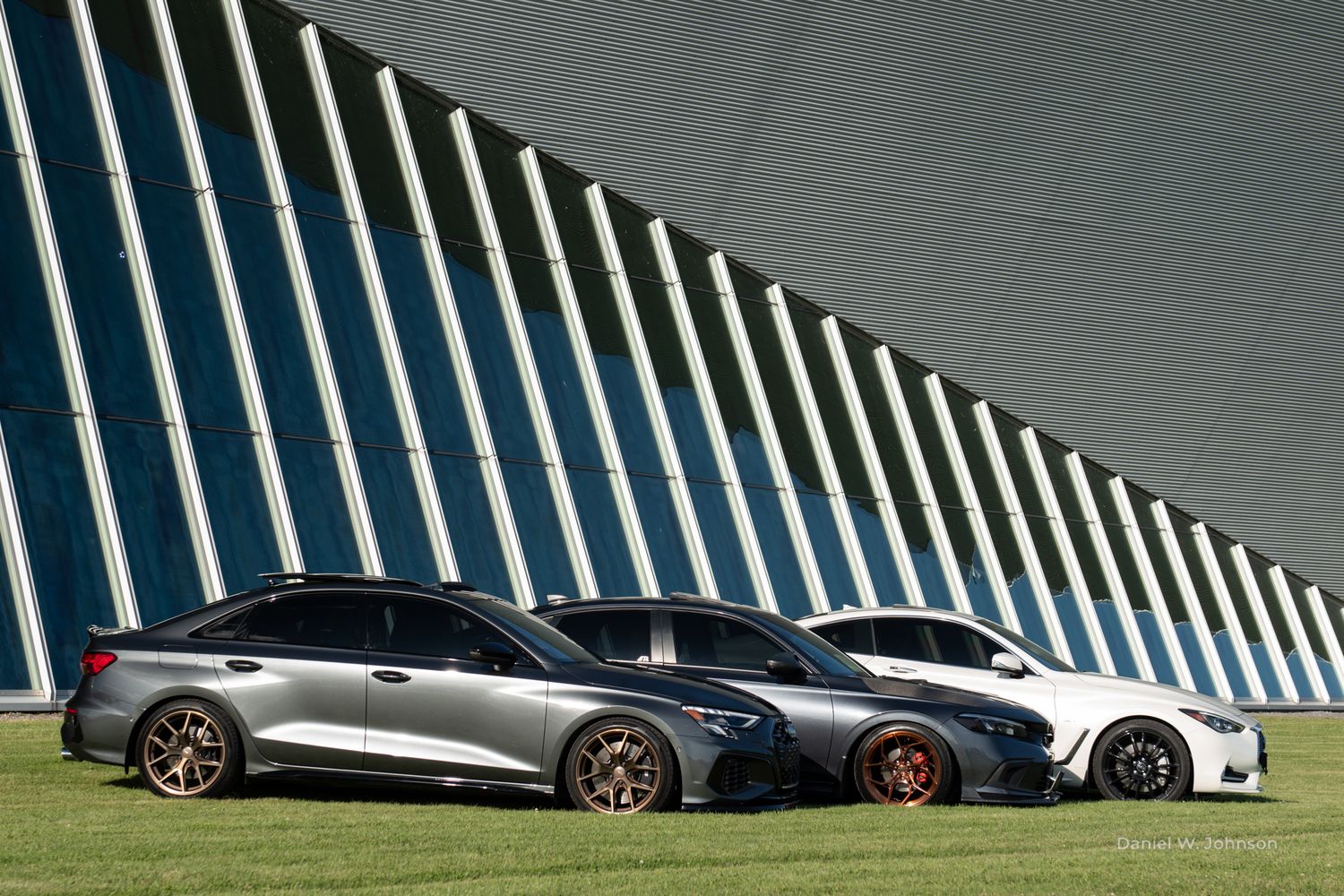 Modern luxury cars parked in a row under an architectural structure with curved striped ceiling.