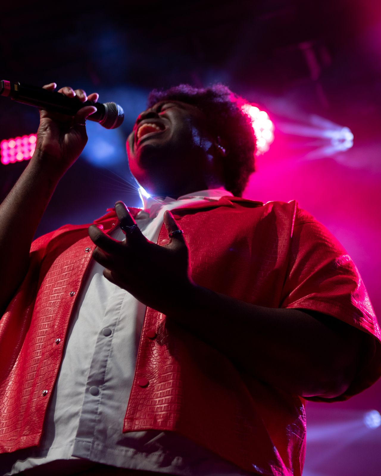 A performer in a red jacket sings passionately on stage with dramatic pink and blue lighting effects in the background.