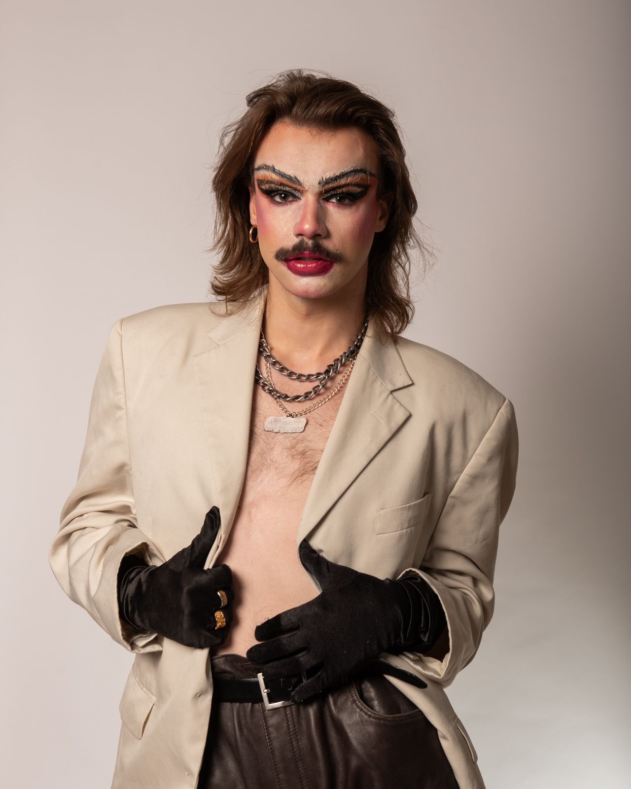 A person in dramatic makeup wearing a beige blazer and black gloves poses for a fashion photoshoot series.