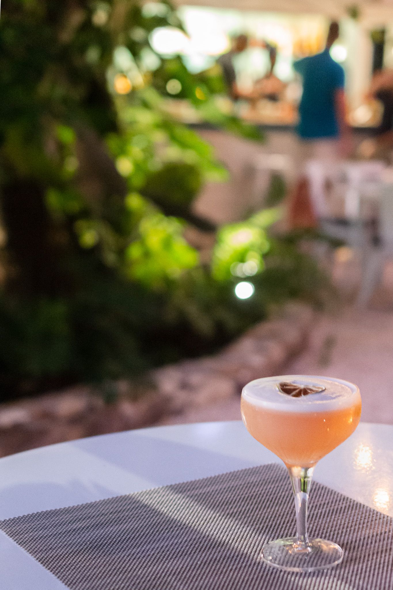 A pink cocktail in a coupe glass sits on a textured gray surface with blurred greenery in the background.