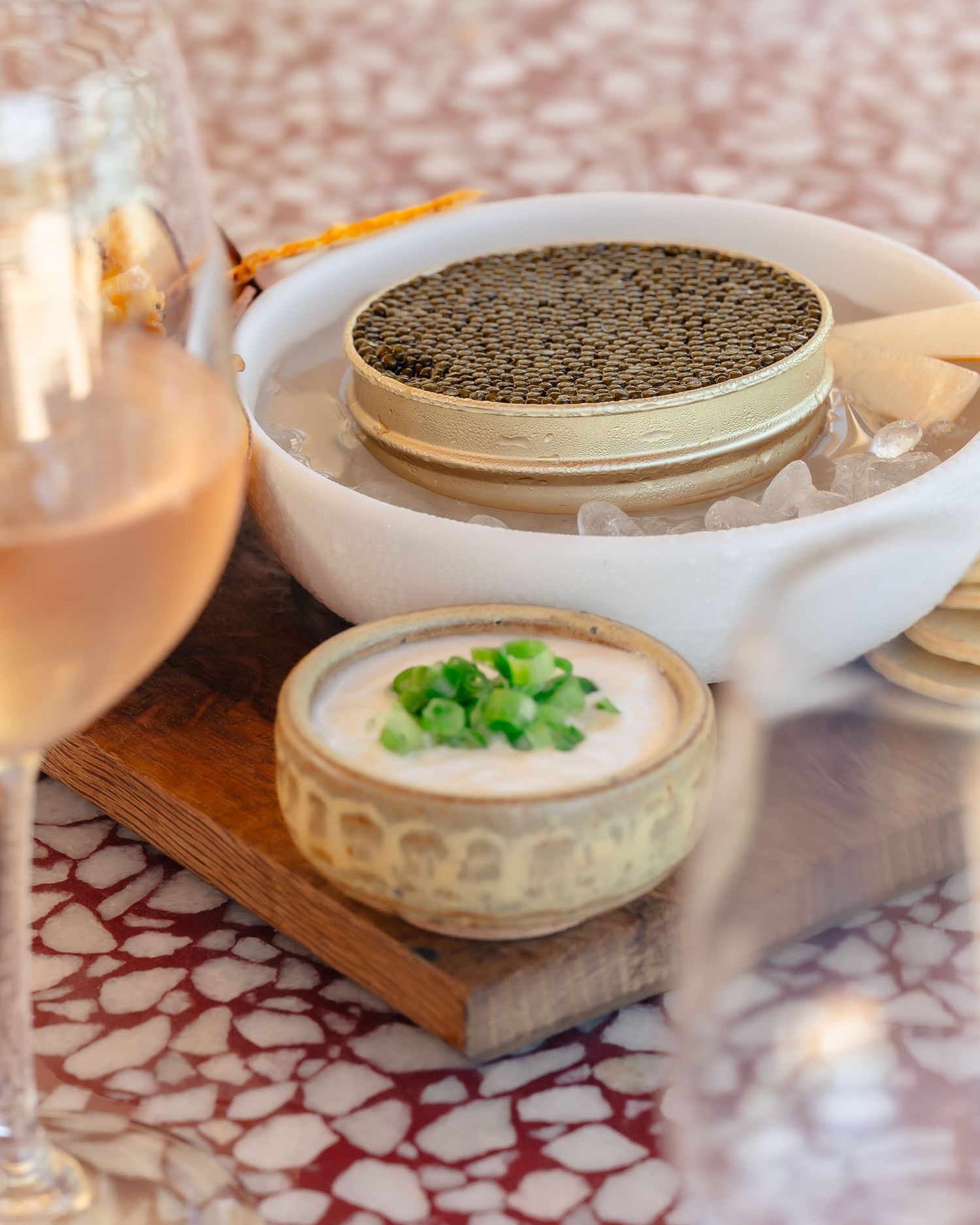 Elegant caviar tin on ice with creamy dip garnished with green onions, served alongside a glass of rosé wine.