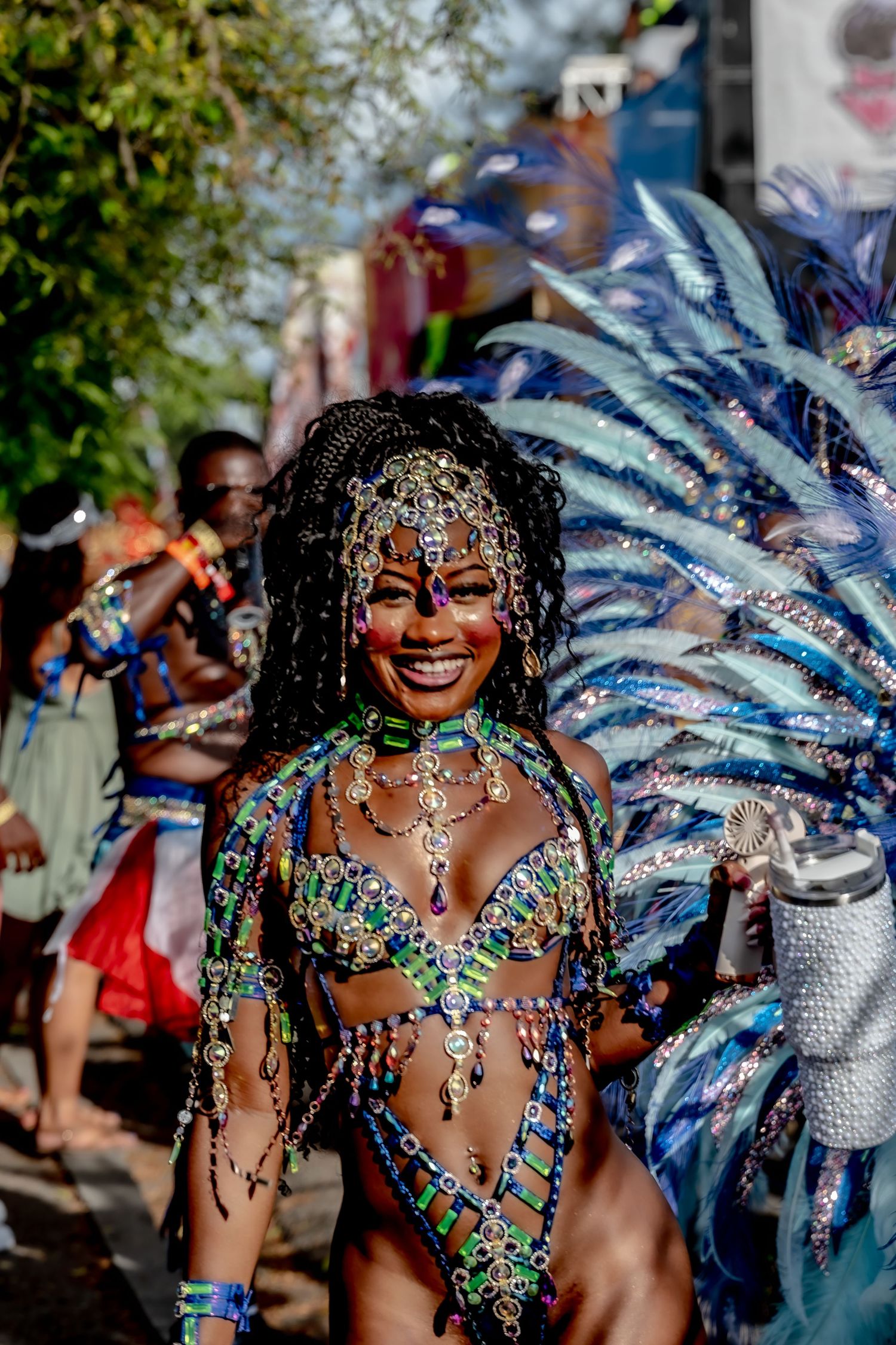 Smiling woman in colorful jeweled carnival costume with blue feathered headdress at a vibrant street festival parade.