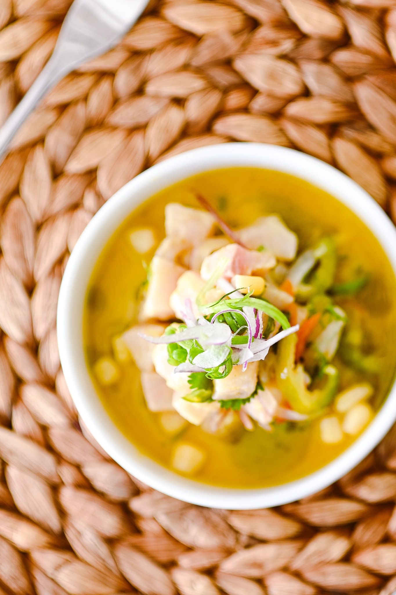 A white bowl of golden soup with chicken, corn, green onions, and herbs on a woven placemat.