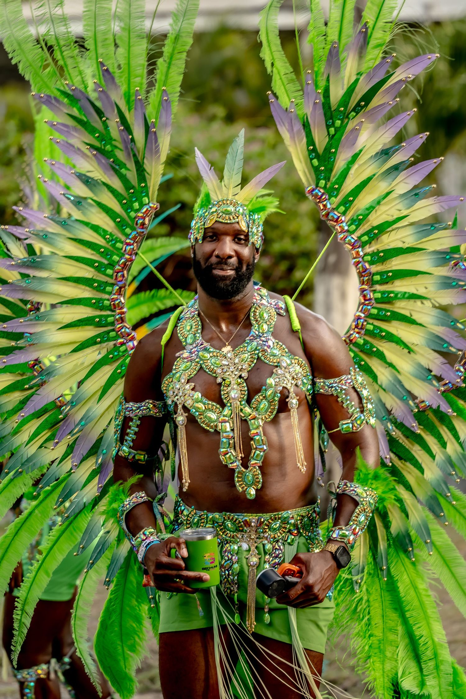 Man in vibrant green feathered carnival costume with jeweled accessories smiling at a festive parade celebration.