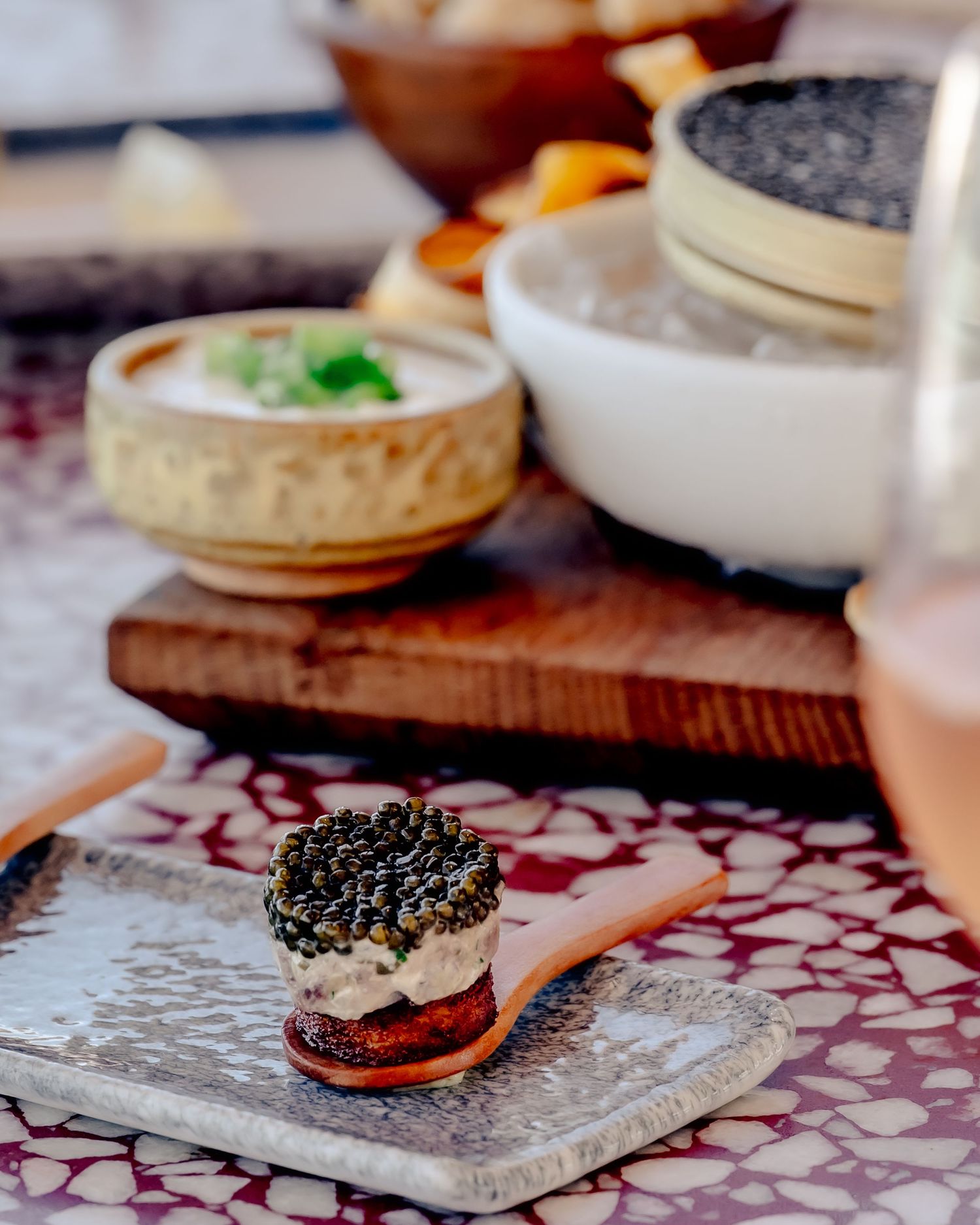 Elegant caviar appetizer on a wooden spoon atop a ceramic plate, with small bowls and a caviar jar in the background.